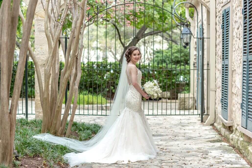 San Antonio wedding photography by Toast: a bride in a lace wedding dress with a long veil holds a bouquet and smiles while standing on a stone path outside near trees and an iron gate.
