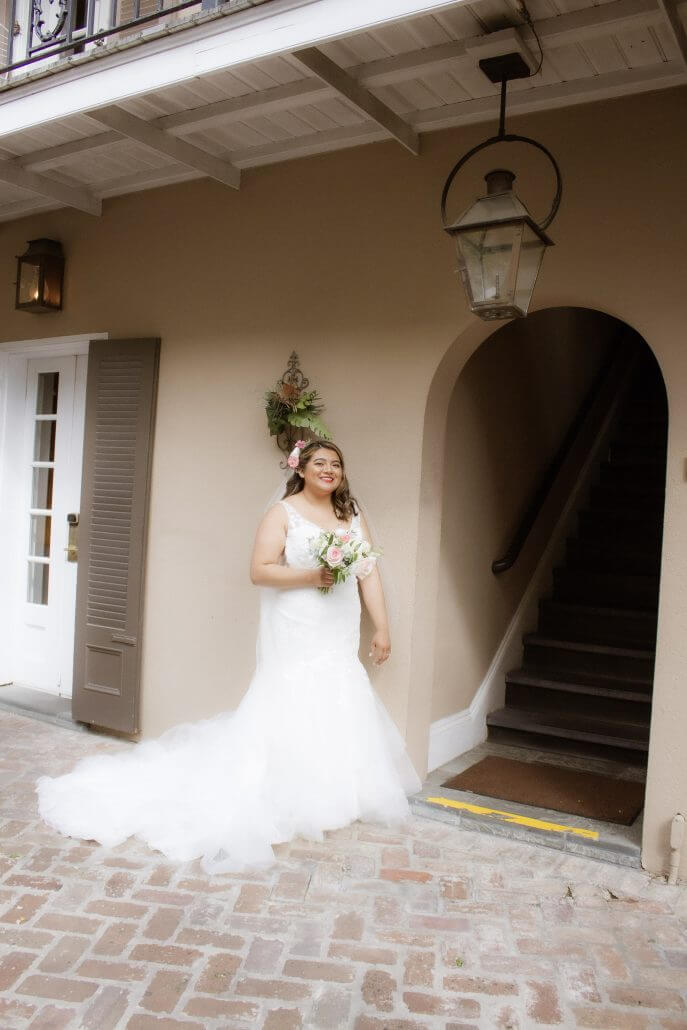 New Orleans wedding photography by Toast: a bride in a white dress holding a bouquet stands outside near a closed door and a staircase, smiling.