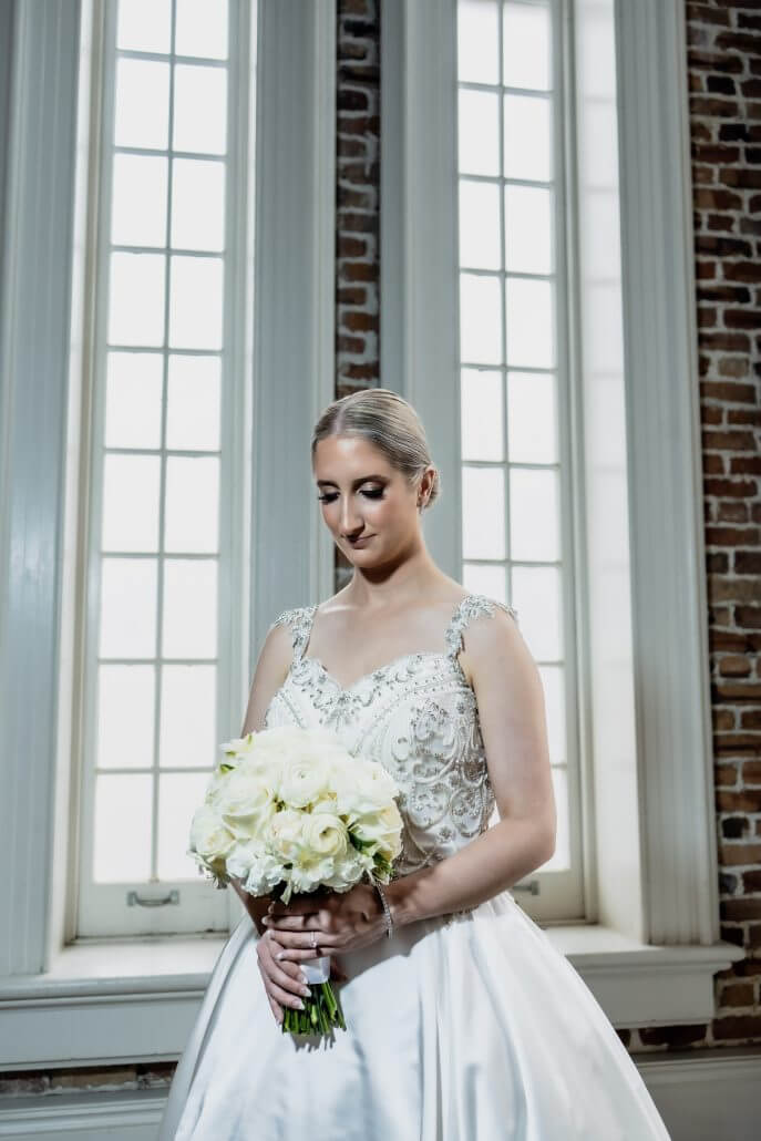 New Orleans wedding photography by Toast: a bride in a white dress holds a bouquet of white roses, standing in front of tall windows with brick walls in the background.