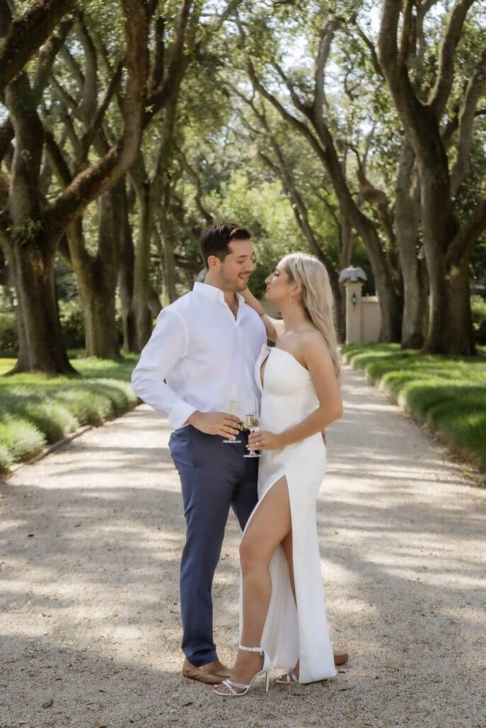 New Orleans wedding photography by Toast: a man and woman stand on a tree-lined gravel path, dressed formally with drinks in hand, looking at each other and smiling.