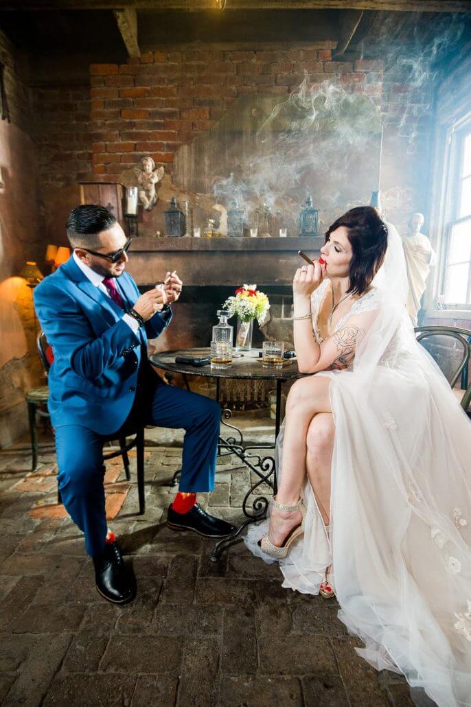 New Orleans wedding photography by Toast: a bride and groom sit indoors at a small table, both lighting cigars; the bride wears a white dress and veil, the groom is in a blue suit, and smoke drifts between them.
