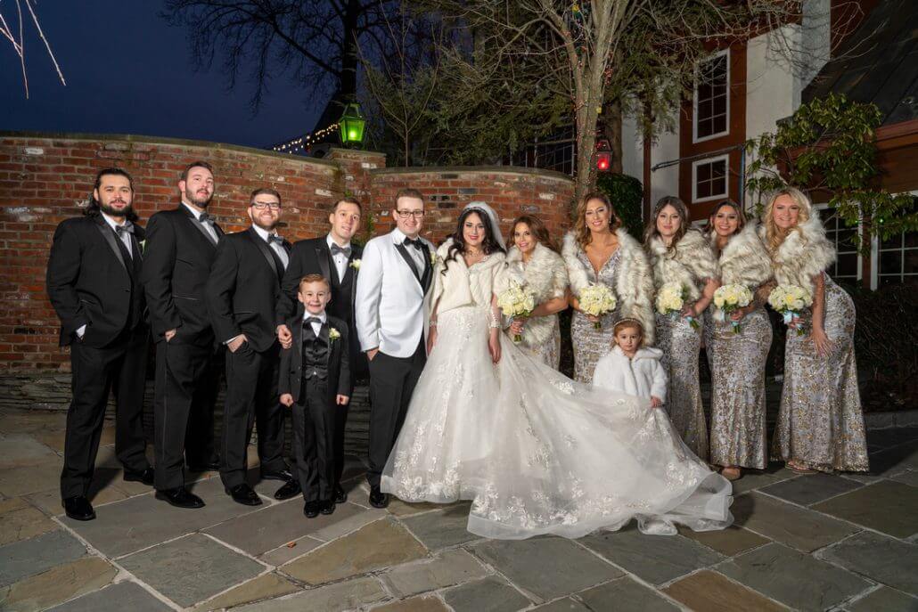 Long Island wedding photography by Toast: a wedding party poses outdoors at night; the bride and groom stand in the center, surrounded by bridesmaids in matching dresses and groomsmen in tuxedos.