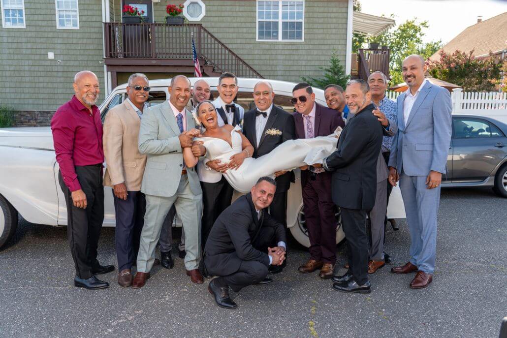 Long Island wedding photography by Toast: a group of men in suits pose outdoors by a vintage car, holding up a smiling woman in a white dress. Everyone is smiling for the photo.