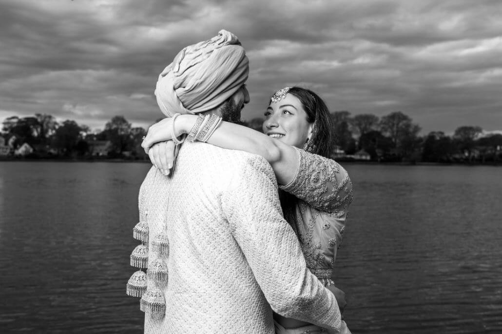 Long Island wedding photography by Toast: a couple dressed in traditional South Asian attire embrace by a lake, with trees and clouds in the background.