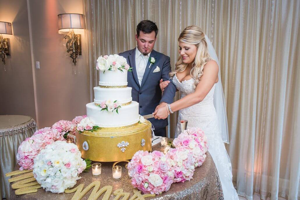 New Orleans wedding photography by Toast: a bride and groom cut a three-tiered white wedding cake together, surrounded by pink and white flower bouquets and candles on a decorated table.