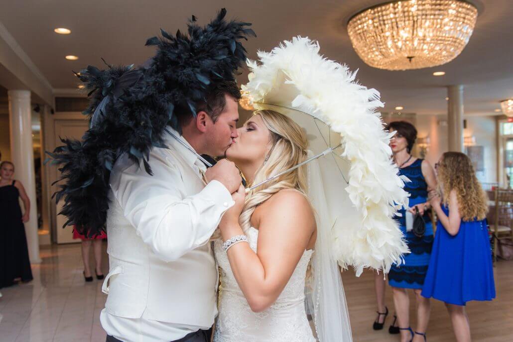 gallery-heather-0022 A bride and groom kiss indoors while holding large feathered hats, with guests standing in the background.