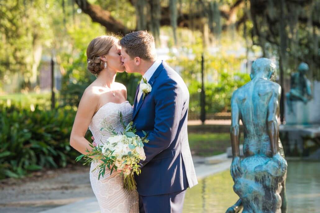 New Orleans wedding photography by Toast: a bride and groom kiss outdoors beside a small reflecting pool and statue, surrounded by greenery and sunlight.