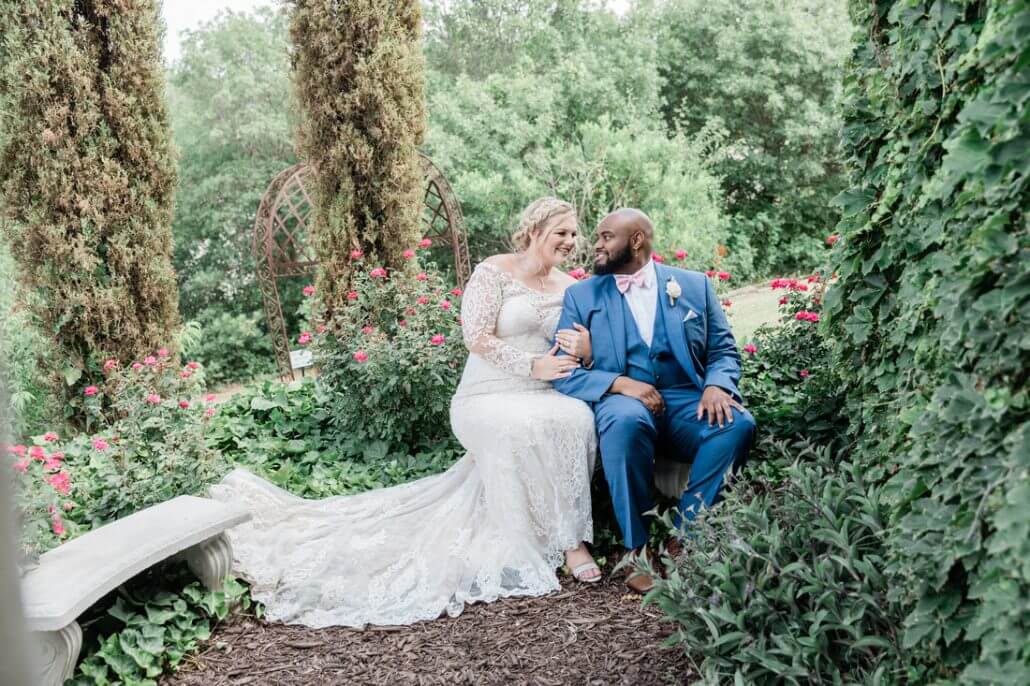 A bride in a white lace dress and a groom in a blue suit sit together on a stone bench in a lush garden, smiling at each other.