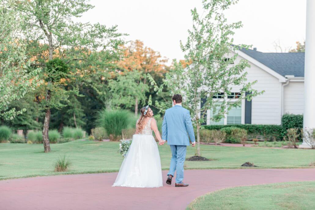 Fort Worth wedding photography by Toast: a bride and groom walk hand in hand on a paved path through a green garden, with a white building visible in the background.