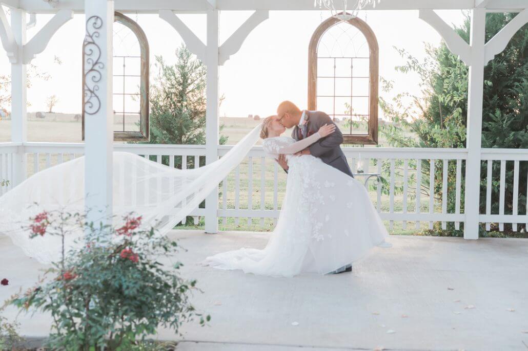 A bride and groom share a kiss while the groom dips the bride under a gazebo; the bride’s veil flows behind her.