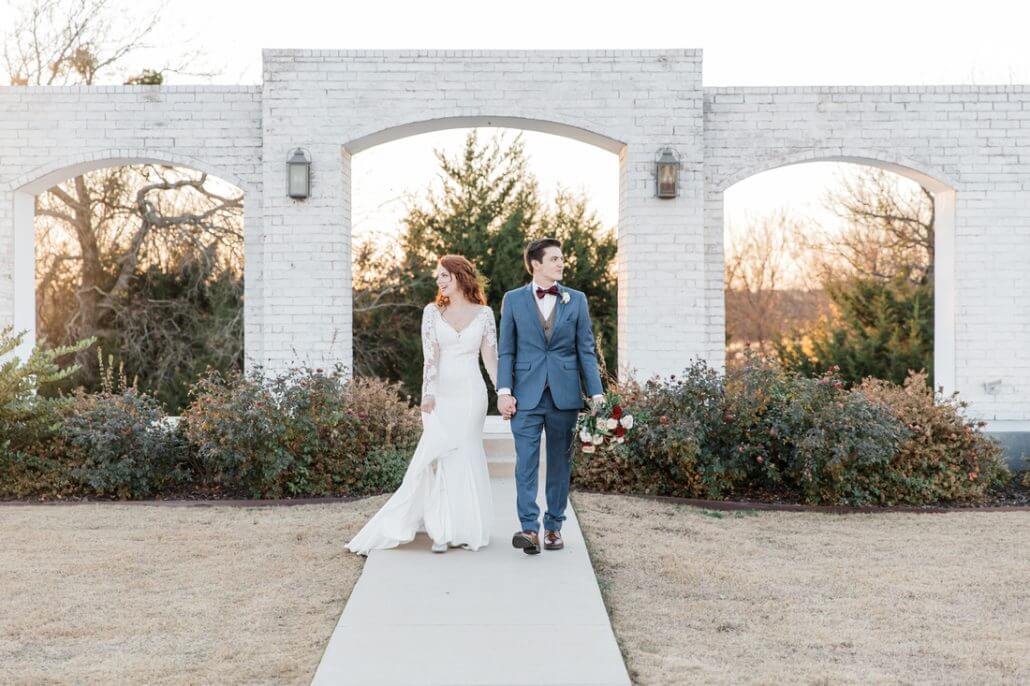 Fort Worth wedding photography by Toast: a bride and groom holding hands and walking outside on a path, with greenery and a white brick structure in the background.