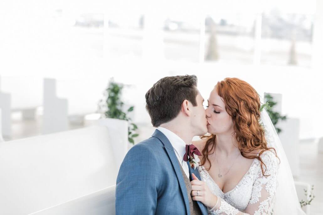A bride and groom sit on a white bench, sharing a kiss. The bride wears a white lace dress and the groom is in a blue suit with a bow tie.