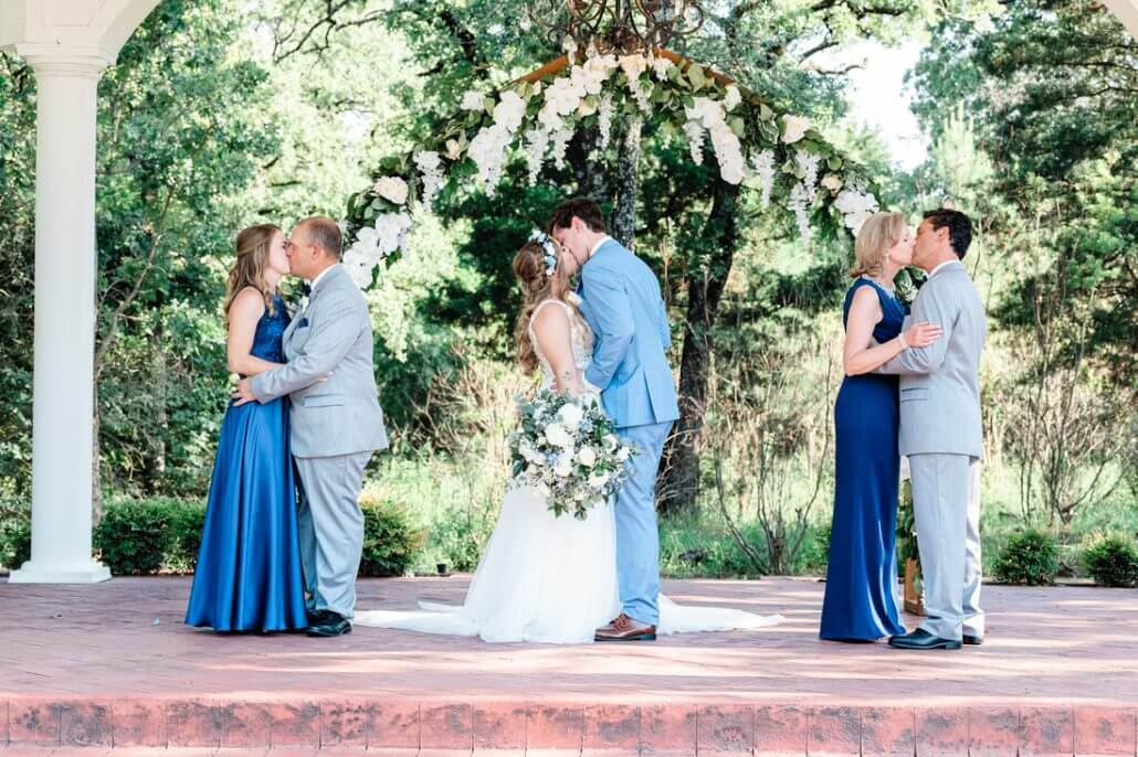 Fort Worth wedding photography by Toast: three couples, dressed in formal attire, stand under a floral arch outdoors. The couple in the center, dressed as bride and groom, kiss, flanked by two other couples also sharing a kiss.