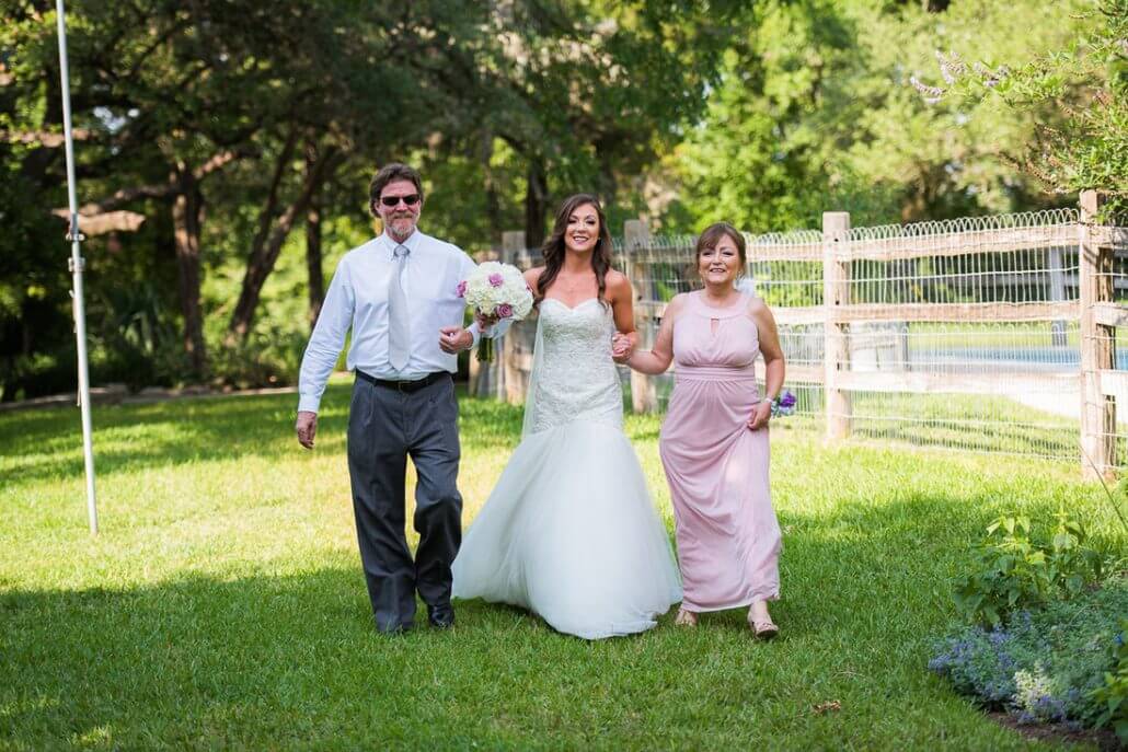 San Antonio wedding photography by Toast: a bride in a white gown walks outdoors on grass, holding flowers, accompanied by a man and a woman in formal attire.