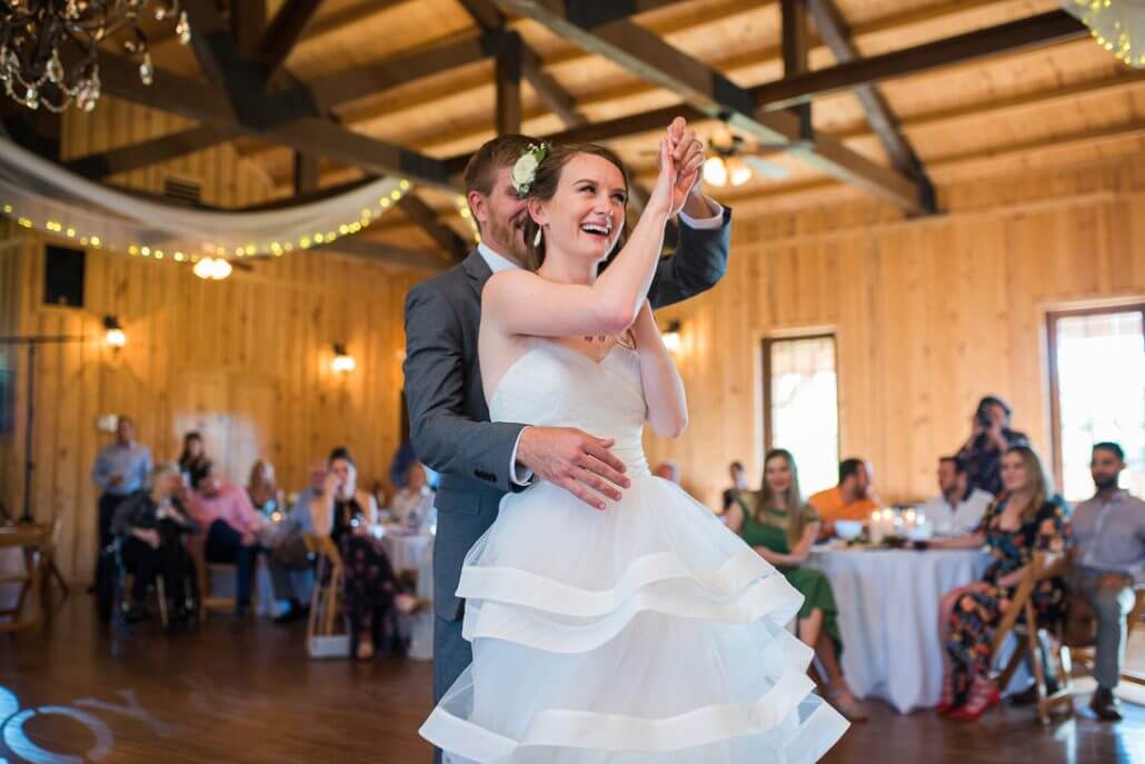 San Antonio wedding photography by Toast: a bride and groom dance together in a wooden hall, smiling and clapping, while guests seated at round tables watch in the background.
