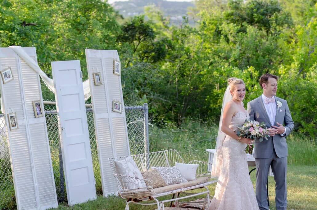 A bride and groom stand outdoors near a bench and white folding screens decorated with framed photos, with greenery in the background.