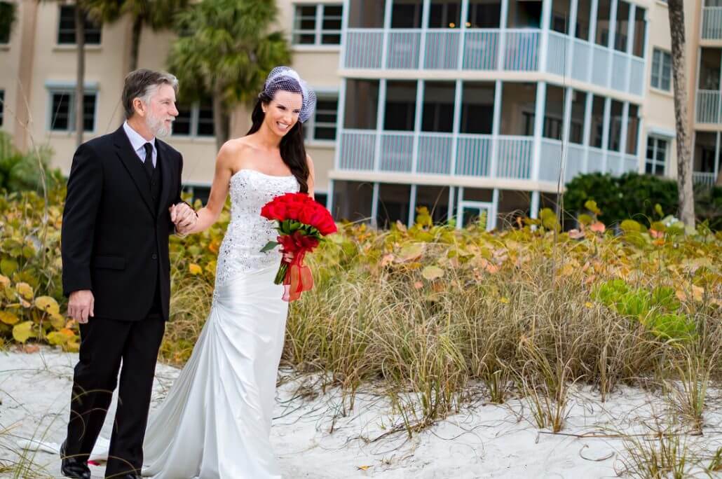 A bride in a white dress holding red roses walks with an older man in a suit on a sandy path, with a building and greenery in the background.