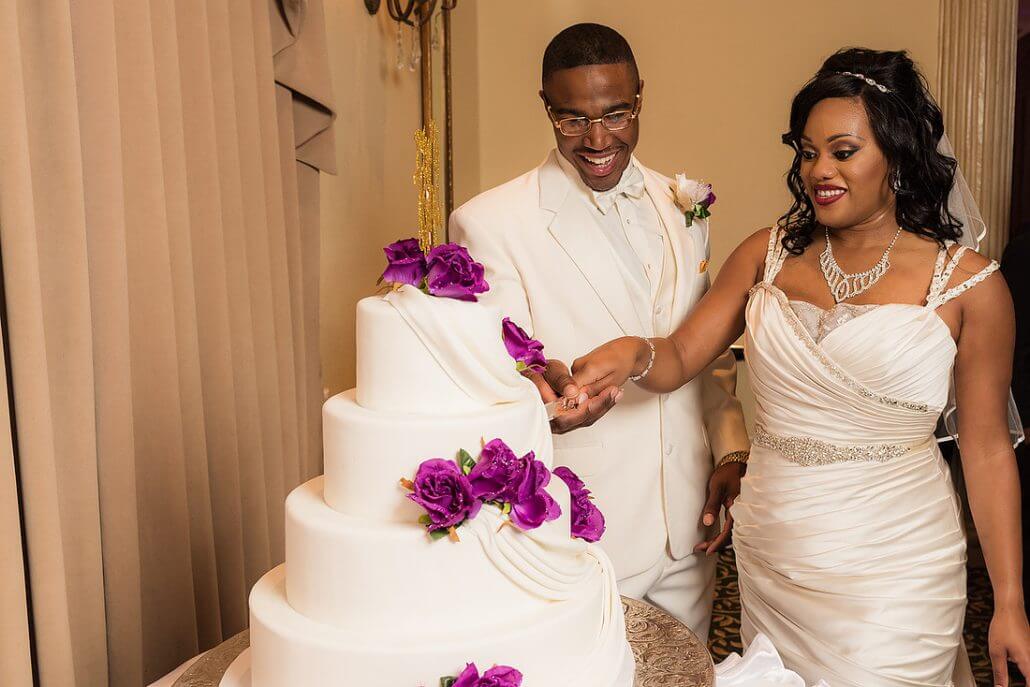 Fort Worth wedding photography by Toast: a bride and groom dressed in white cut a tiered wedding cake decorated with purple flowers.