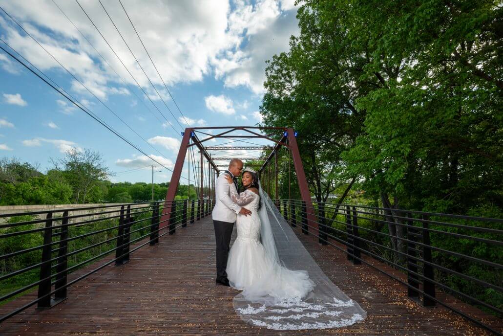 Fort Worth wedding photography by Toast: a bride and groom stand embracing on a wooden bridge, surrounded by greenery, with the bride's veil flowing behind her under a partly cloudy sky.