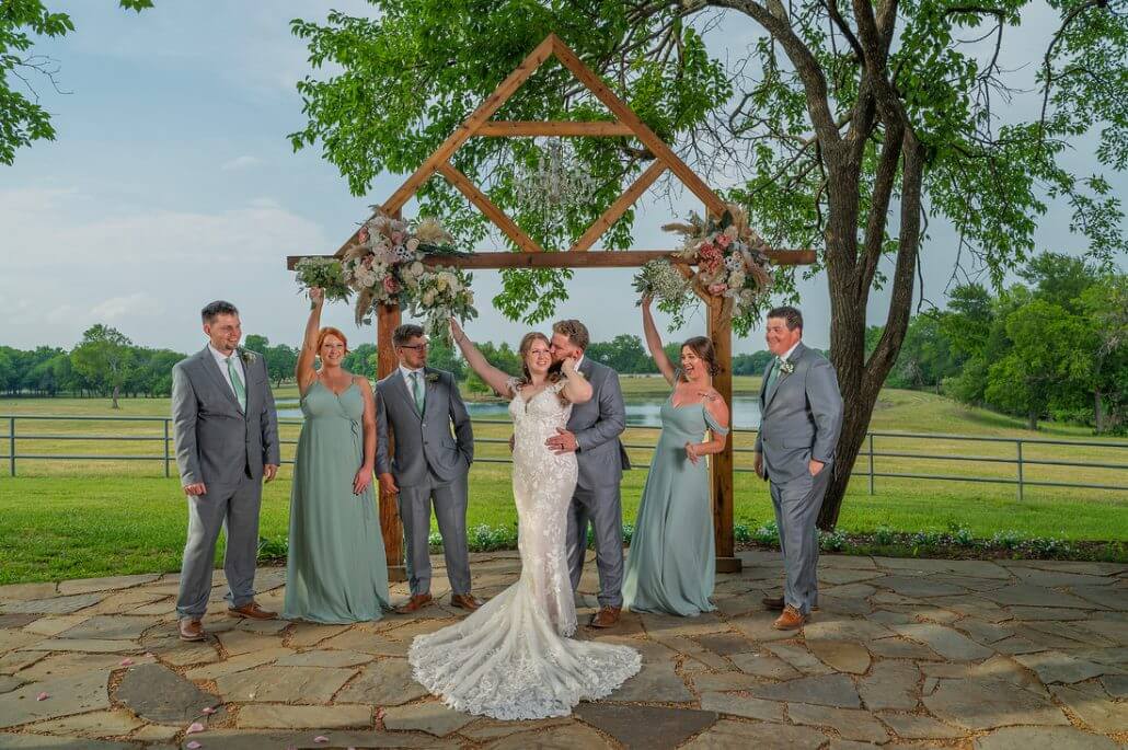 Fort Worth wedding photography by Toast: a bride and groom stand under a wooden wedding arch with four attendants in matching gray suits and green dresses, posing outdoors on a stone patio with grassy fields in the background.