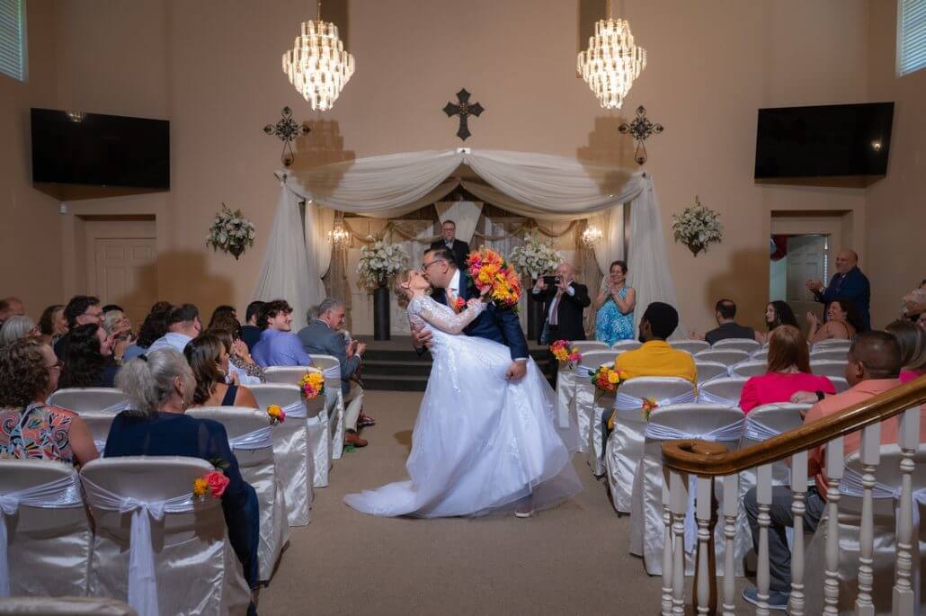 A bride and groom kiss at the altar during a wedding ceremony in a decorated indoor venue, surrounded by seated guests.