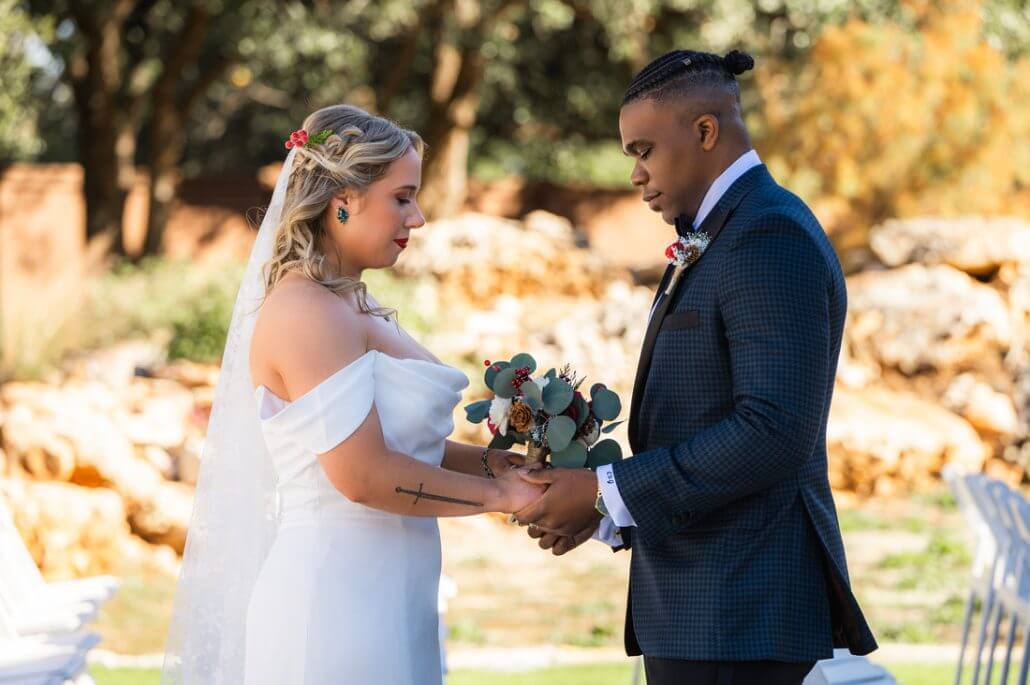 A bride and groom stand facing each other outdoors, holding hands and a bouquet, during their wedding ceremony.