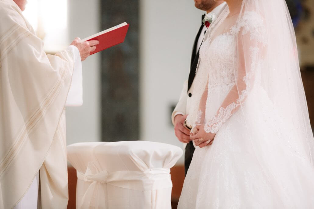 A bride and groom stand at the altar during a wedding ceremony, facing an officiant holding an open book.