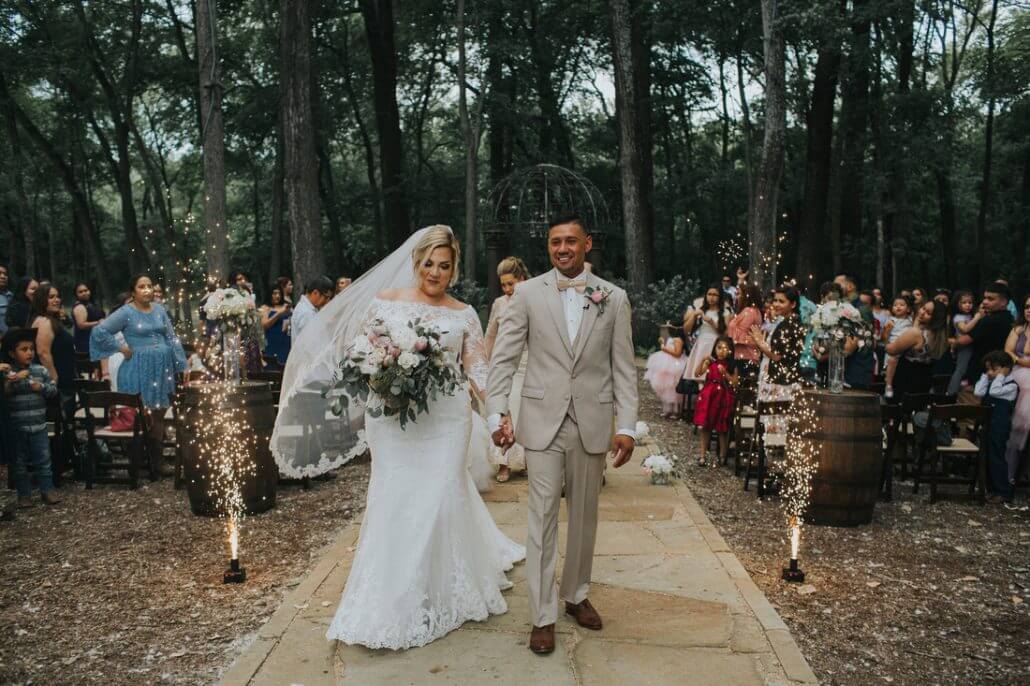 gallery-victor-0076 Bride and groom walk down an outdoor aisle lined with guests and sparklers, surrounded by trees on their wedding day.