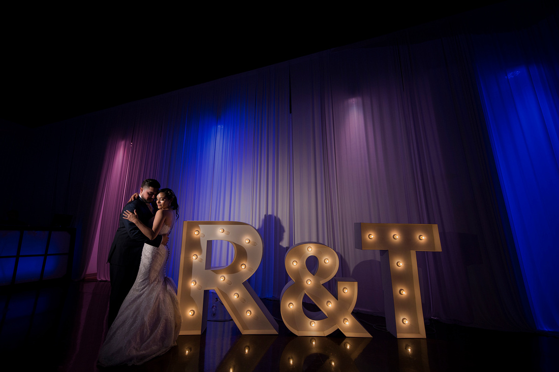 gallery-victor-0232 A bride and groom embrace on a dimly lit dance floor next to large illuminated letters "R & T," with purple and blue lighting in the background.