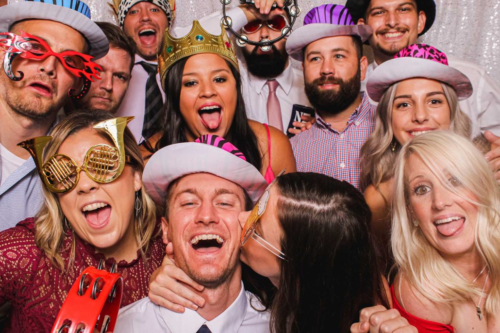 toast-entertainment-photo-booth-group A group of people wearing hats and novelty glasses pose closely together, smiling and laughing, in front of a glittery backdrop.