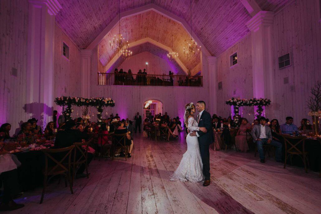A bride and groom share their first dance in a large, warmly lit hall with guests seated at decorated tables and purple accent lighting.