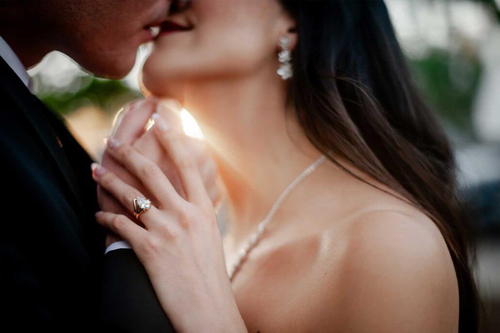 Close-up of a couple kissing, with the woman wearing a diamond ring and pearl necklace, and sunlight shining between them.