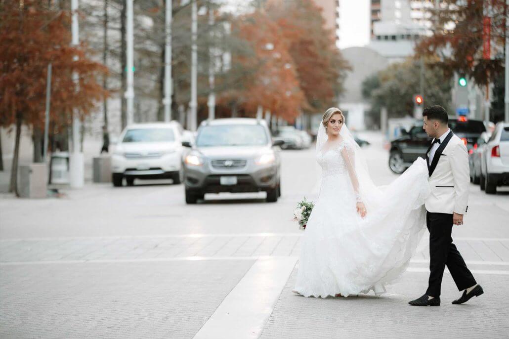 Bride and groom crossing a downtown Dallas street on their wedding day while the groom carries her train and traffic waits.
