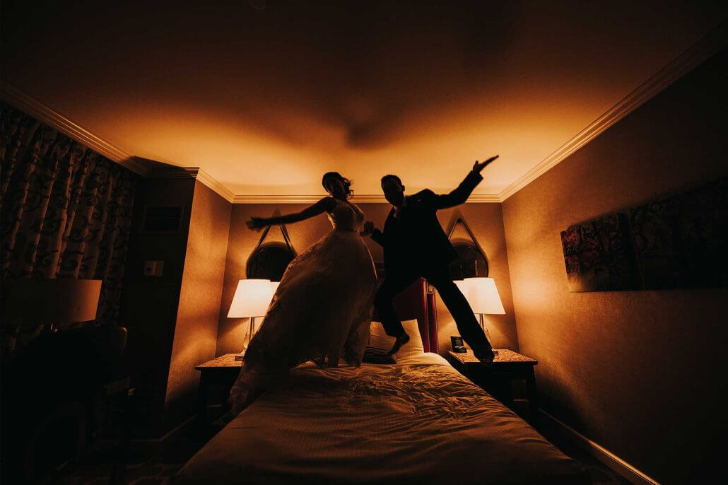 Fort Worth wedding photography by Toast: silhouettes of a bride and groom jumping on a bed in a dimly lit hotel room, with warm lighting from lamps on either side.