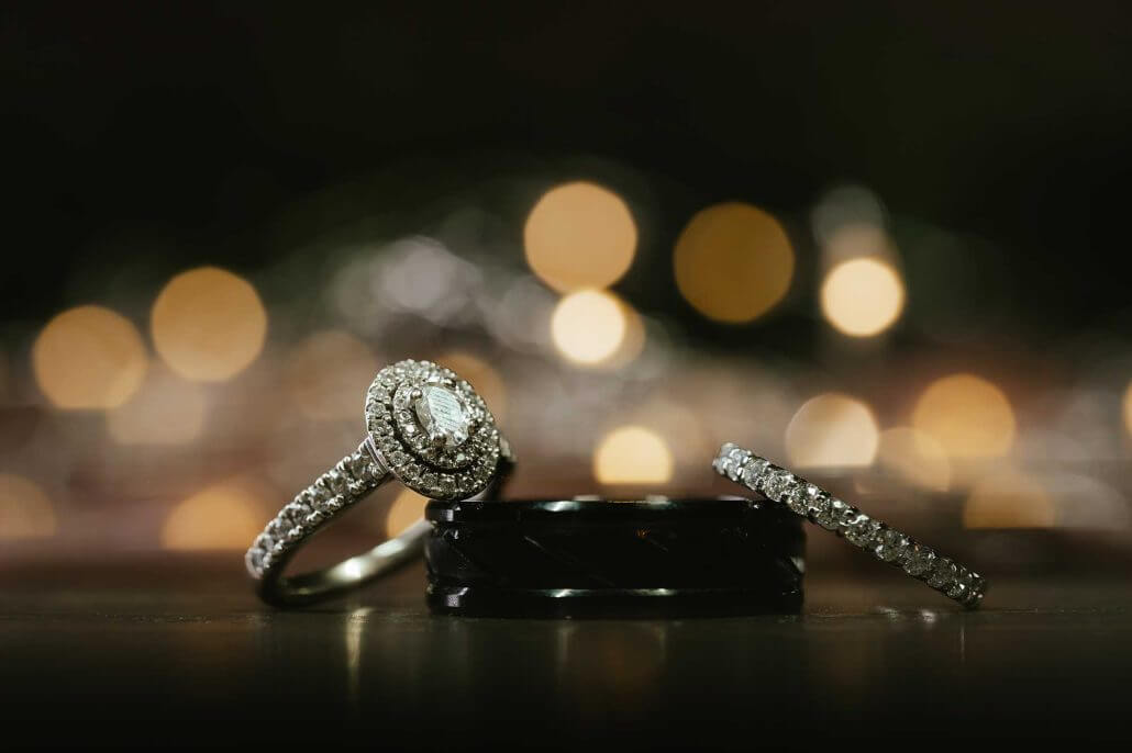 Fort Worth wedding photography by Toast: three rings, including a diamond engagement ring, a wedding band with diamonds, and a black band, are displayed on a surface with blurred lights in the background.
