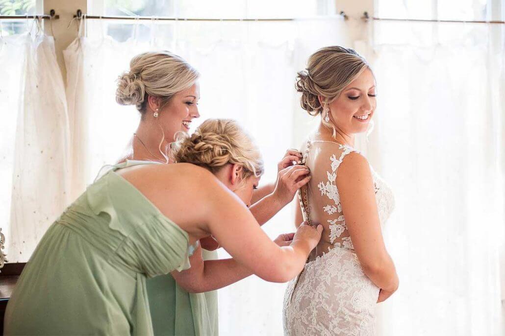 Houston wedding photography by Toast: two women help a bride in a lace wedding dress fasten the back of her gown as she stands smiling in a softly lit room with white curtains.