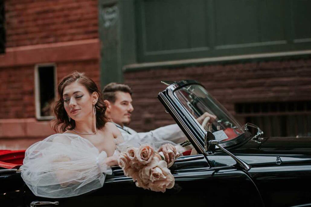 Houston wedding photography by Toast: a bride holding a bouquet sits in the passenger seat of a black convertible with a man driving, both in formal attire, on a city street.