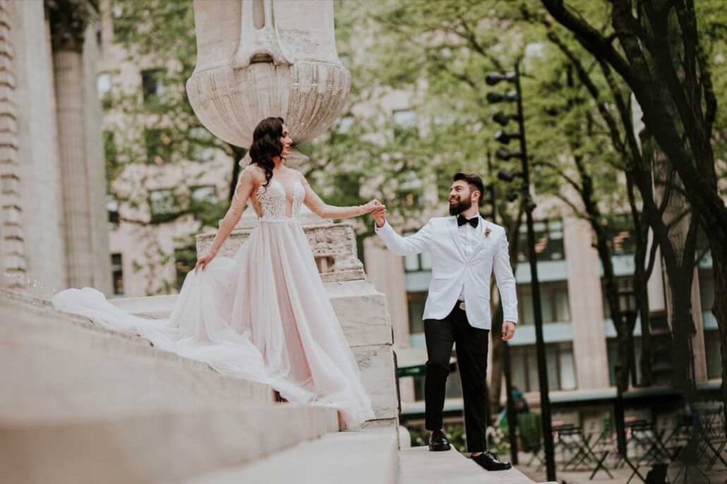 Houston wedding photography by Toast: a bride in a light pink dress and a groom in a white tuxedo walk down stone steps outdoors, holding hands, with trees and buildings in the background.