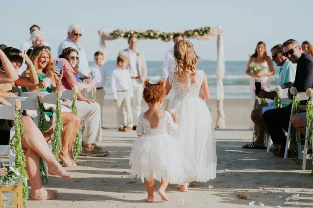 Houston wedding photography by Toast: two young flower girls in white dresses walk down the aisle at an outdoor beach wedding, with guests seated on either side and the ocean visible in the background.