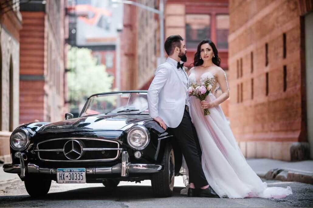 Bride and groom posing beside a black vintage Mercedes convertible on a brick-lined street, from a Toast Long Island wedding.