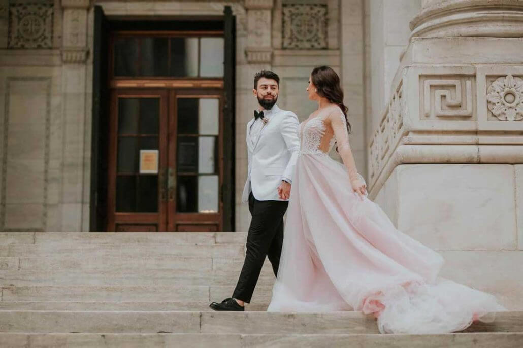 Long Island wedding photography by Toast: a couple dressed in formal attire, with the woman in a flowing light pink gown and the man in a white jacket, walk up stone steps outside an ornate building.