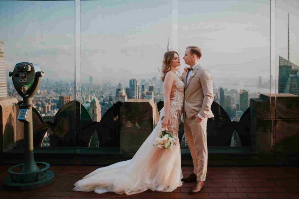 Long Island wedding photography by Toast: a bride and groom stand together on a rooftop terrace overlooking a city skyline at sunset, with a coin-operated viewer nearby.