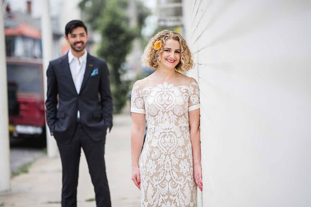New Orleans wedding photography by Toast: a woman in a white lace dress stands by a white wall, smiling, with a man in a dark suit and open collar shirt standing in the background on a sidewalk.