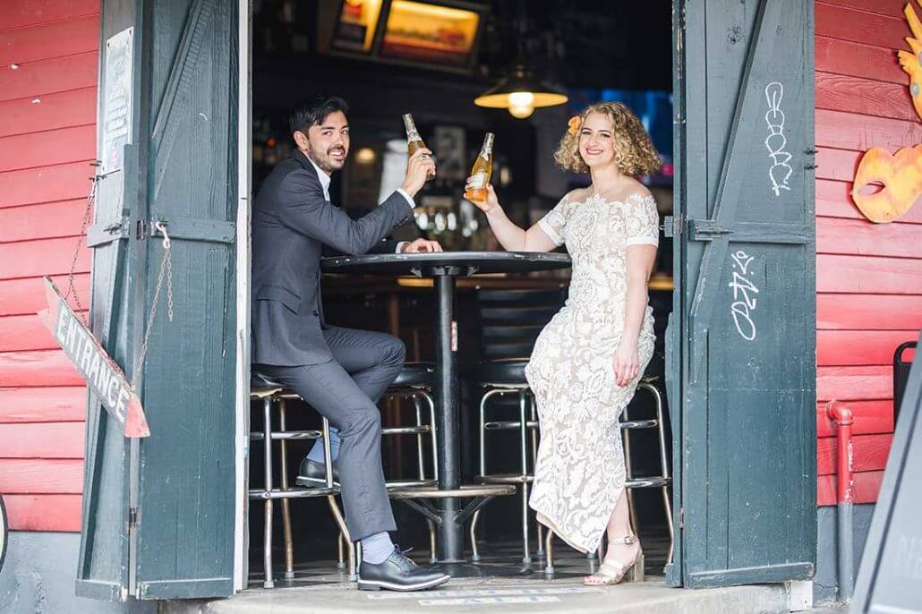 New Orleans wedding photography by Toast: a man in a suit and a woman in a white dress sit at a bar table inside an open doorway, clinking beer bottles and smiling.