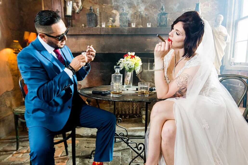 New Orleans wedding photography by Toast: a man in a blue suit and a woman in a wedding dress sit at a table, both smoking cigars, with drinks and flowers on the table in a dimly lit room.