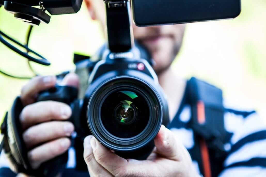 photography videography A person holding a DSLR camera with a mounted microphone and screen, wearing a striped shirt and camera harness, is aiming the lens toward the viewer.