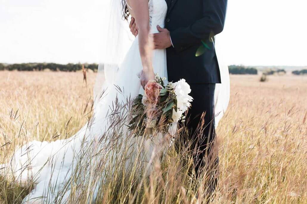 South Texas wedding photography by Toast: a bride and groom stand close together in a field of tall grass; the bride holds a bouquet of flowers and wears a white dress.