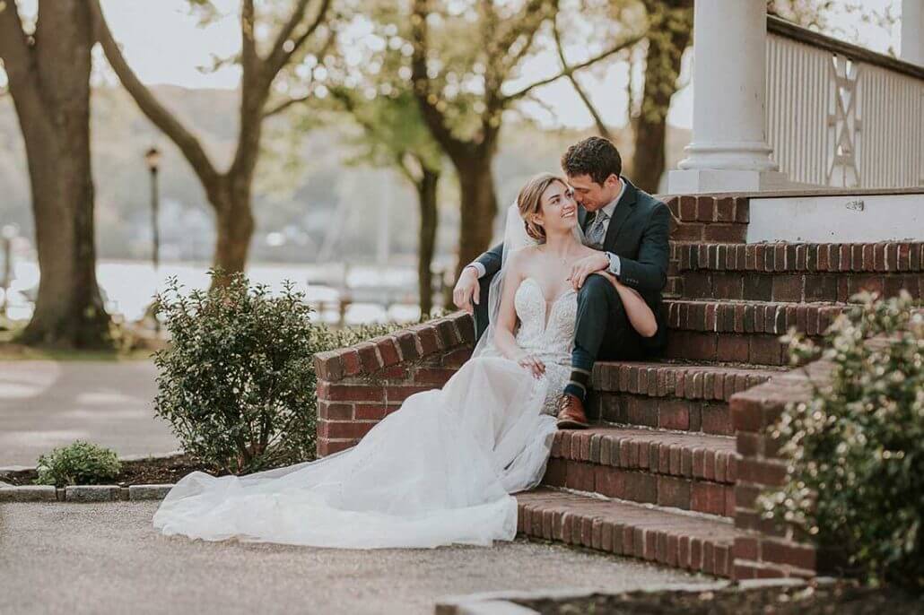 South Texas wedding photography by Toast: a bride in a white gown and a groom in a dark suit sit closely together on outdoor brick steps, surrounded by greenery, on a sunny day.