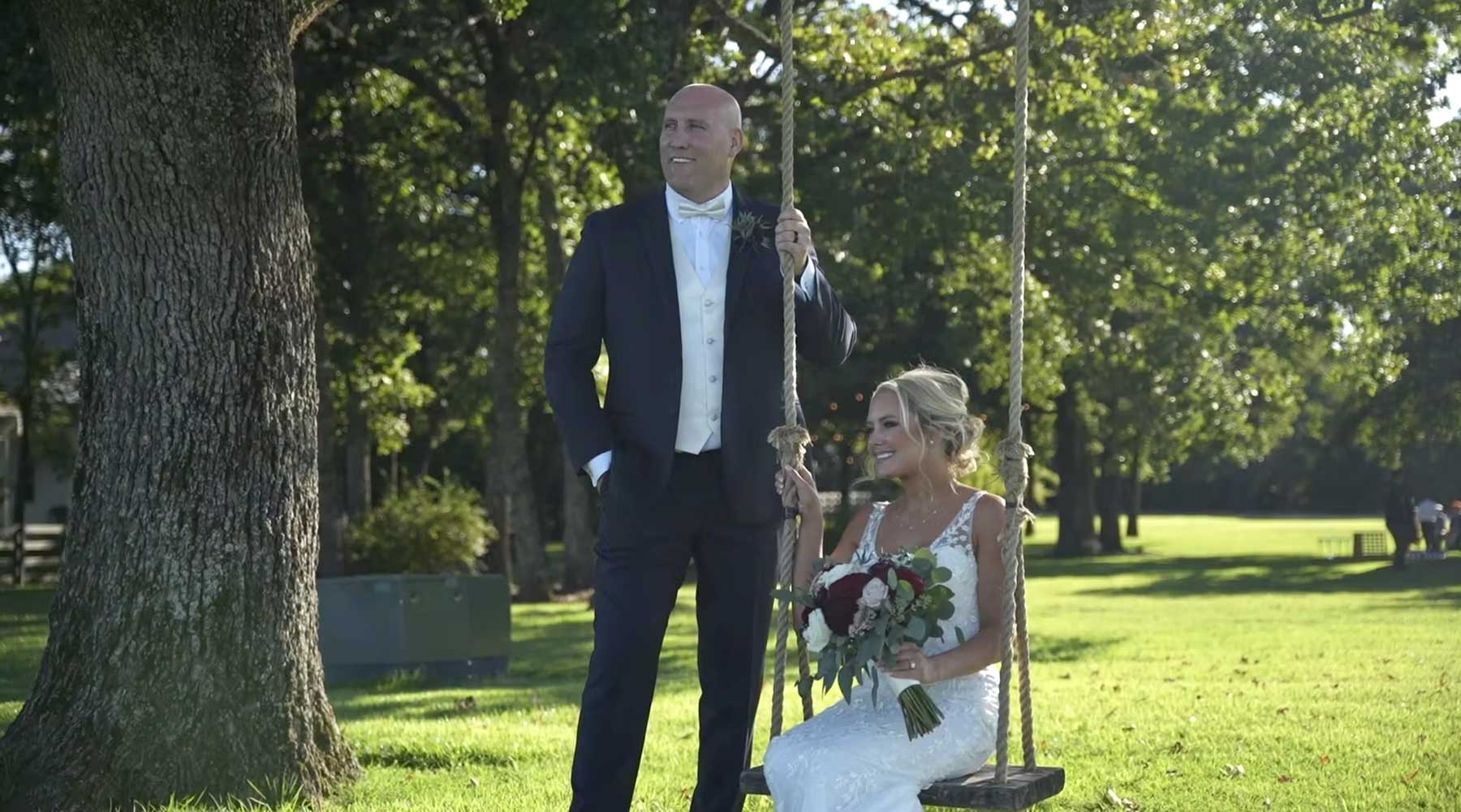 A bride in a white dress holding a bouquet sits on a swing while a groom in a tuxedo stands beside her outdoors on a sunny day.