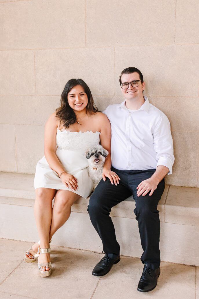 A woman in a white dress, a man in a white shirt and black pants, and a small dog sit together on a stone bench in front of a beige wall.